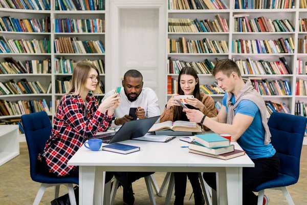 Vue de face de quatre sympathiques étudiants multiethniques souriants de 25 ans utilisant leur smartphone pour les réseaux sociaux pendant la pause pendant leur apprentissage ou leur tâche à la maison dans la bibliothèque moderne . — Photo