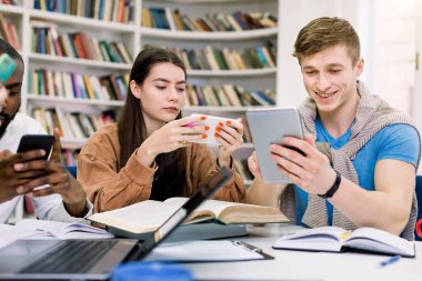 Çok ırklı arkadaşlar, üniversite öğrencileri kütüphanede okuyor ve sınavlara hazırlanırken aletlerini, akıllı telefonlarını ve tabletlerini kullanıyorlar. Kitaplar, dizüstü bilgisayarlar ve defterler masanın üstünde.