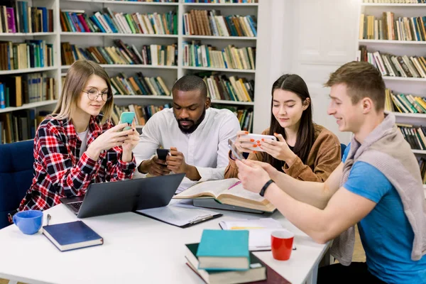 Quatre jeunes, deux garçons et deux filles étudiants, assis à table, travaillant ensemble, à l'aide de leurs téléphones intelligents, ordinateur portable et livres. Groupe multiracial d'étudiants faisant des études de groupe dans une bibliothèque . — Photo