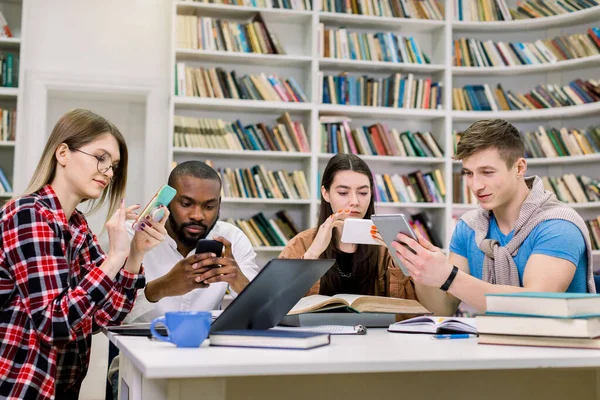 Cuatro jóvenes amigos estudiantes, niños y niñas de raza mixta, que se están preparando para los exámenes y la búsqueda de la información necesaria en sus teléfonos inteligentes, sentado en la sala de lectura en la biblioteca — Foto de Stock