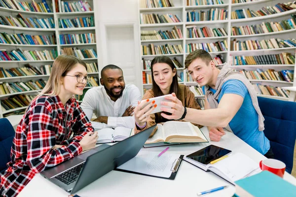 행복해 보이는 네 명의 친구들, 행복해 보이는 혼합 인종 학생들 이 책꽂이에 있는 도서관에서 그룹 연구를 하는 동안웃긴 비디오에서 스마트폰을 보고 있습니다. — 스톡 사진