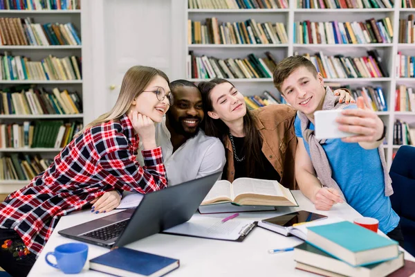 Multiétnicos sonrientes estudiantes felices están estudiando en la biblioteca, divirtiéndose durante el descanso y haciendo foto selfie. Los jóvenes están pasando tiempo juntos, sentados con libros y portátiles en la biblioteca . — Foto de Stock