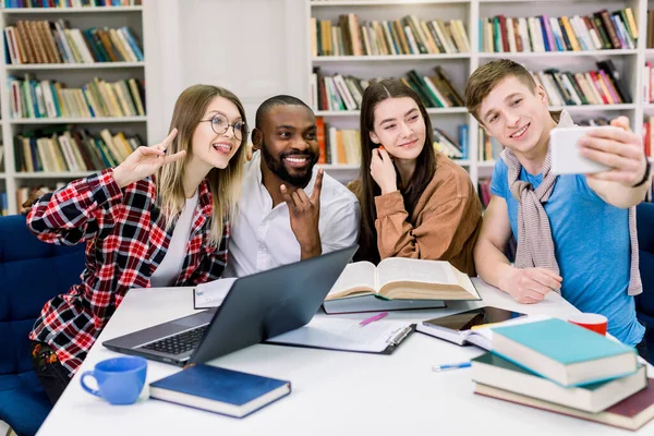 Jovens estudantes universitários felizes fazendo selfie foto juntoresr enquanto estudam na biblioteca. Grupo de pessoas multirraciais na biblioteca da faculdade sentados juntos à mesa com livros e laptop — Fotografia de Stock