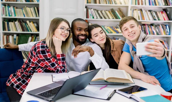 Personas, diversión, educación, tecnología y el concepto de la escuela - felices cuatro estudiantes multiétnicos haciendo foto divertida selfie en la biblioteca, mientras estudian juntos. Educación divertida, academia, escuela — Foto de Stock