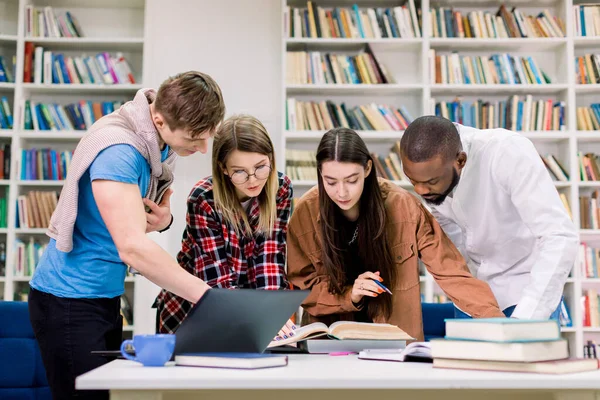 Front view of four concentrated multiethnic young people, college students preparing to their homework in library room, looking for needed information in book. Laptop and books on the table