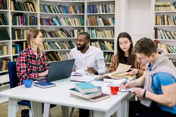Cuatro estudiantes felices multirraciales sentados a la mesa en la biblioteca mientras aprenden y trabajan en una computadora portátil. Estudiantes inteligentes que trabajan juntos en la sala de lectura usando libros y computadoras portátiles — Foto de Stock