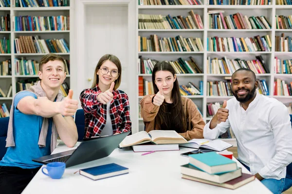 Alegre sorrindo quatro estudantes 25s multirraciais, dois meninos e duas meninas, sentados à mesa com livros e laptop na sala de leitura da biblioteca, olhando para a câmera e mostrando os polegares sinal ok para cima — Fotografia de Stock