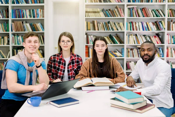 Retrato de jovem quatro grupo estudantil multirracial sentado à mesa com livros e laptop, estudando juntos na biblioteca da universidade, olhando para a câmera e sorrindo — Fotografia de Stock