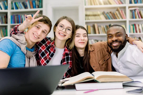 大学生们坐在桌旁，看着镜头，开心地微笑着，互相拥抱着。桌上的书和笔记本电脑。在图书馆阅览室一起学习的年轻人 — 图库照片