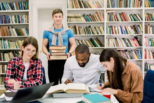 Multietnická skupina mladých lidí, studentů, kteří studují společně u stolu, čtou knihy. Mladý chlapec drží hromadu knih stojí za stolem a dívá se na své koncentrované přátele — Stock fotografie
