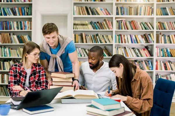 Bonito estudante jovem satisfeito colocando muitos livros diferentes sobre a mesa para seus amigos multi-raciais da universidade, sentado e estudando juntos na moderna sala de leitura da biblioteca do campus . — Fotografia de Stock