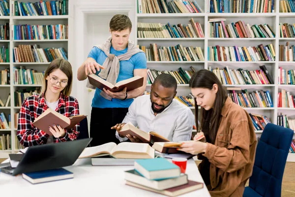 Grupo internacional de quatro jovens estudantes inteligentes focados em roupas casuais que estudam na biblioteca universitária moderna, sentados à mesa com livros e laptop, lendo livros para obter informações — Fotografia de Stock