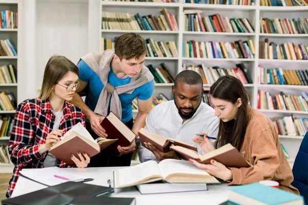 Inteligentes jóvenes estudiantes multiétnicos confiados leyendo libros en la mesa, buscando información y estudiando juntos en la biblioteca de la universidad —  Fotos de Stock