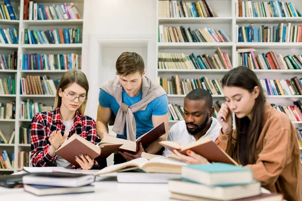 Cuatro amigos multiétnicos concentrados inteligentes sentados a la mesa en la biblioteca universitaria y leyendo libros juntos, preparándose para el examen o la clase, haciendo tareas en casa o buscando información —  Fotos de Stock