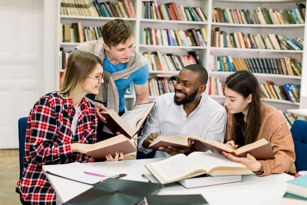 Молоді багатоетнічні друзі, дві жінки і два чоловіки, розмовляють один з одним і посміхаються, читаючи книги за столом в сучасній бібліотеці в кампусі. Білі книжкові полиці на фоні — стокове фото