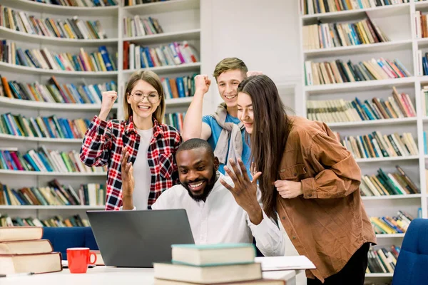 Fröhlich aufgeregte junge multiethnische Freunde feiern erfolgreiche Prüfungsergebnisse oder beenden ihr Projekt, schauen auf den Bildschirm des Laptops, in der modernen Campus-Bibliothek oder im Coworking-Café — Stockfoto