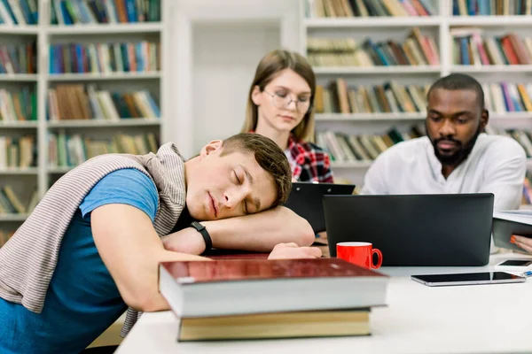 Κοντινό πλάνο του κουρασμένος ύπνο όμορφος μαθητής αρσενικό βρίσκεται στα χέρια του και τα βιβλία στο τραπέζι. Συγκεντρωμένοι μαθητές πολλών εθνοτήτων που προετοιμάζονται για τις εξετάσεις, καθισμένοι στο τραπέζι στο παρασκήνιο — Φωτογραφία Αρχείου