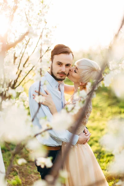 Щаслива пара в елегантному розкішному одязі, гуляє в красивому квітучому саду, насолоджуючись весняною природою. Красивий чоловік дивиться на камеру і посміхається, поки його гарненька жінка цілує щоку — стокове фото