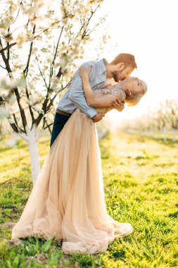 Güzel, neşeli ve romantik bir çift çiçek açan bahçede poz veriyor. Zarif elbiseli güzel kadınının boynunu öpen yakışıklı bir adam büyük bir tutku ve aşkla. Bahar düğün portresi