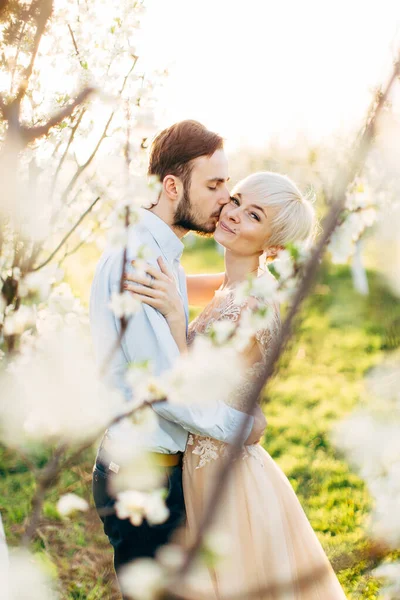 Щаслива пара в елегантному розкішному одязі, гуляє в красивому квітучому саду, насолоджуючись весняною природою. Красива жінка дивиться на камеру і посміхається, поки її красивий чоловік цілує її щоку — стокове фото