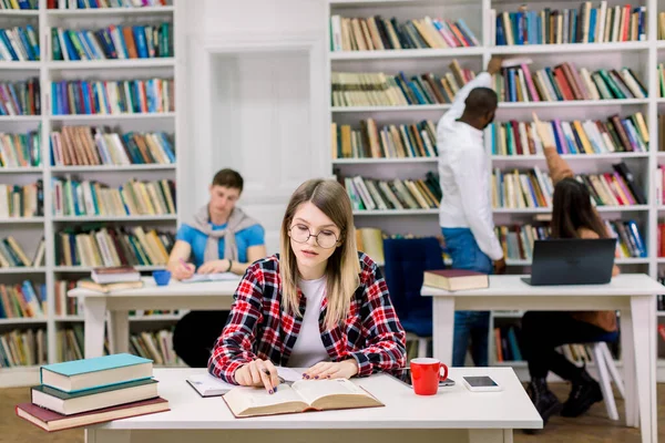 大学図書館の読書室で試験の準備をしながら、本からノートを書いて、眼鏡や小切手シャツで魅力的なスマート自信のある現代の女の子。背景には多民族 — ストック写真