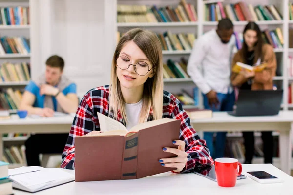 Jovem 25 anos caucasiana bela estudante menina em óculos estudando, lendo informações necessárias do livro enquanto sentado à mesa na biblioteca da universidade moderna. Estudantes de raça mista em segundo plano — Fotografia de Stock