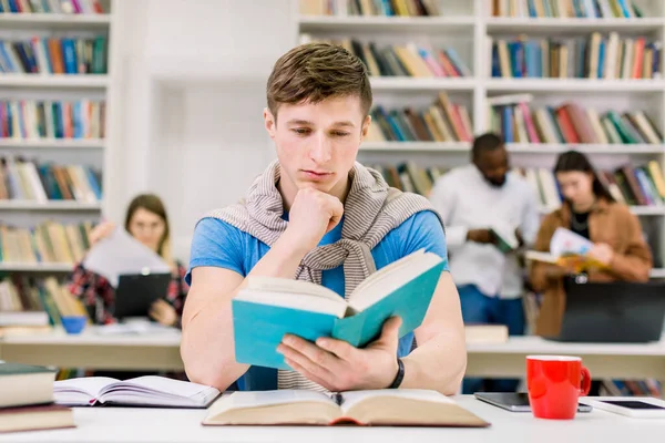 Esperto confiante jovem caucasiano que estuda na biblioteca para o teste ou exame, ele está sentado na mesa e lendo um livro, enquanto seus colegas de grupo discutindo sobre o fundo. Conceito de conhecimento e aprendizagem — Fotografia de Stock