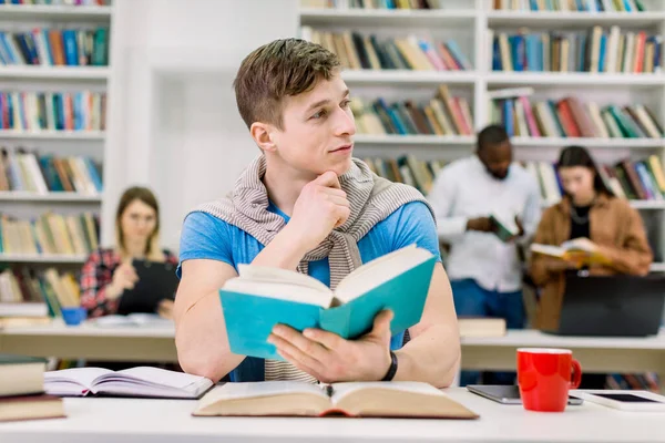 Pensamento atraente bonito estudante do sexo masculino sentado à mesa com muitos livros e se preparando para o teste ou exame. Estudantes multiétnicos trabalhando juntos e biblioteca interior em segundo plano — Fotografia de Stock