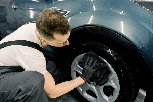 Vista lateral del apuesto trabajador de lavado de autos caucásico aullando en paño de microfibra negro de la mano y limpiando la rueda del coche. Lavado de coches y concepto de detalle — Foto de Stock