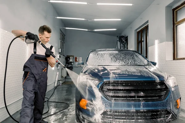Zastřelen mladým bělochem, který myje modré auto pod vysokým tlakem vody v myčce aut. Odstranění mýdla vodou, čištění auta — Stock fotografie