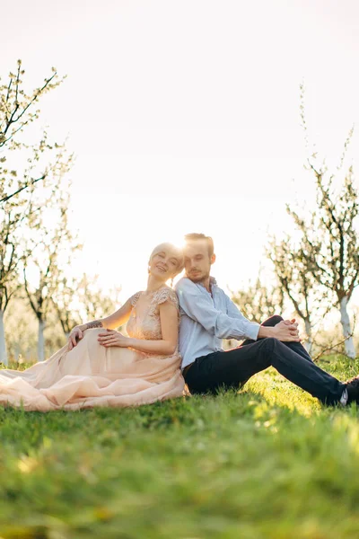 Close up retrato do jovem casal caucasiano apaixonado, vestindo roupas de casamento de luxo, sentado de volta para trás na grama verde no belo jardim florescente primavera. Primavera retrato de casamento ao ar livre — Fotografia de Stock