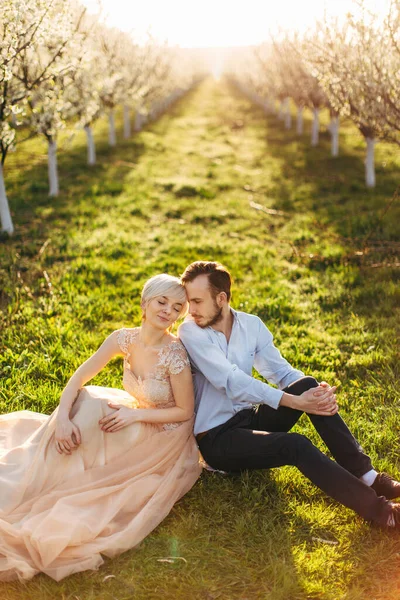 Jovem casal encantador sentado de volta para trás na grama verde no fundo do belo nascer do sol e árvores floridas no jardim. Mulher bonita em vestido longo, apoiando-se em seu belo homem barbudo — Fotografia de Stock