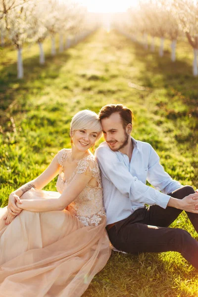 Jovem belo casal romântico no jardim contra o fundo de cerejeiras, sentados na grama e inclinando-se para trás uns aos outros — Fotografia de Stock