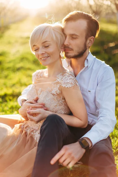 Casal jovem no jardim florescente primavera ter data romântica, sentado na grama. Mulher loira bonita em vestido laranja claro, apoiando-se em seu belo homem barbudo e sorrindo — Fotografia de Stock