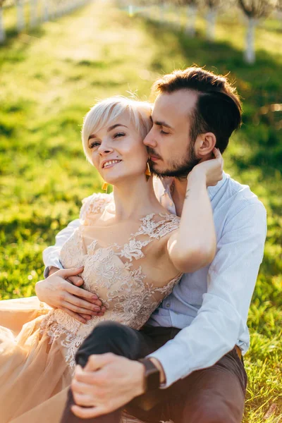 Casal jovem no jardim florescente primavera ter data romântica, sentado na grama. Mulher loira bonita em vestido laranja claro, apoiando-se em seu belo homem barbudo e sorrindo — Fotografia de Stock