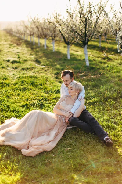 Feliz pareja caucásica romántica agradable sentado en la hierba verde y abrazos, en el hermoso jardín en flor, disfrutando de la naturaleza de primavera. Concepto de ternura y amor — Foto de Stock