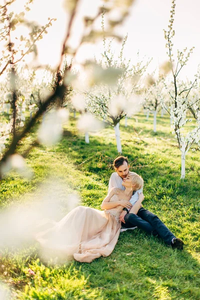 Joyeux couple amoureux profitant d'une journée ensoleillée dans le jardin fleuri du printemps. Jolie femme en robe longue s'appuyant sur son amant et relaxant. Vue à travers la branche fleurie — Photo