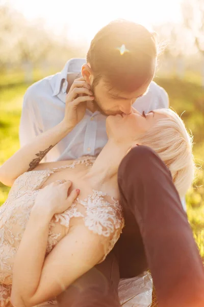Chrapanie młodej blondynki w eleganckiej sukience leżącej na kolanach swojego chłopaka i całującej go. Portret romantycznej czułej pary siedzącej na trawie w wiosennym ogrodzie, słoneczny poranek, wschód słońca — Zdjęcie stockowe