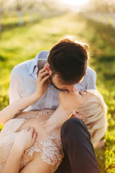 Chraming jovem loira mulher em vestido elegante deitado de joelhos em seu namorado e beijando-o. Retrato de casal terno romântico sentado na grama no jardim da primavera, manhã ensolarada, nascer do sol — Fotografia de Stock