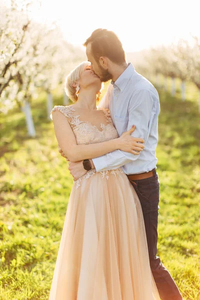 Молода красива поцілункова весільна пара в саду на тлі квітучих дерев. Жінка в розкішній довгій сукні цілує свого красивого коханця. Портрет напівдовжини — стокове фото