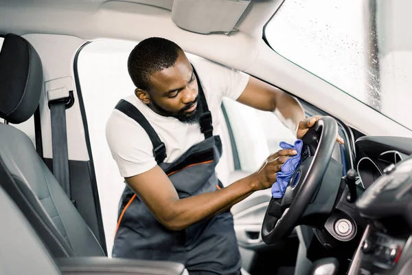 Servicio de limpieza, concepto de detalle del coche. El hombre joven africano en el uniforme de trabajo lava el interior del automóvil, el volante del coche con la ropa azul de microfibra, en el lavado profesional del automóvil — Foto de Stock