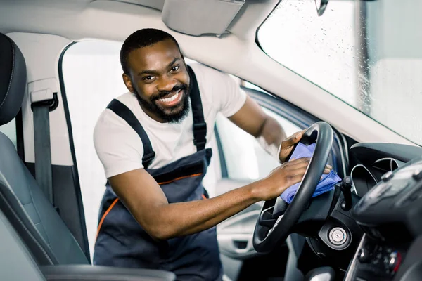 Mikrofiber ve arabanın içini temizleme ve temizleme. Neşeli gülümseyen Afrikalı erkek işçi iç mekan temizleme modern araba, mavi mikrofiber giysiler ve temizlik solüsyonu ile araba direksiyonu — Stok fotoğraf