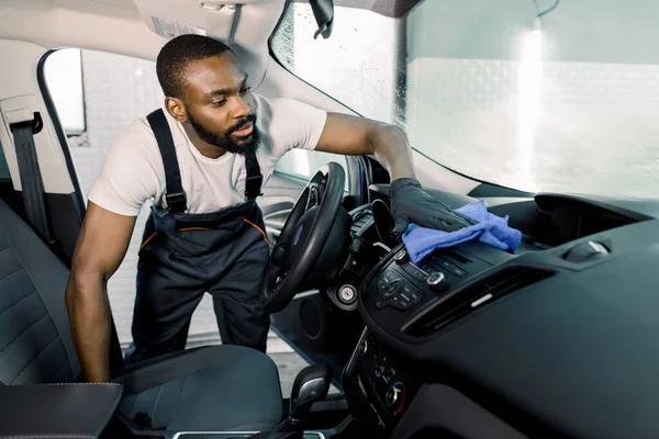 Joven trabajador profesional africano, con monos de trabajo y guantes de goma, limpieza del interior del coche con un paño de microfibra azul — Foto de Stock