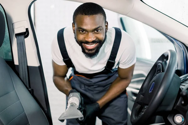 Young handsome African man cleaning car front seat with wet vacuum cleaner, looking at camera and smiling. Car wet chemical vacuum cleaning, car detailing concept