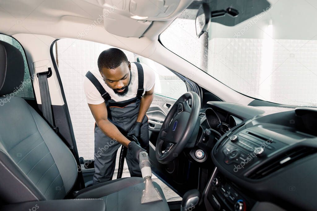 Car interior detailing concept. Young African American man in uniform doing chemical cleaning of gray leather seats with professionally extraction method, using wet vacuum cleaner