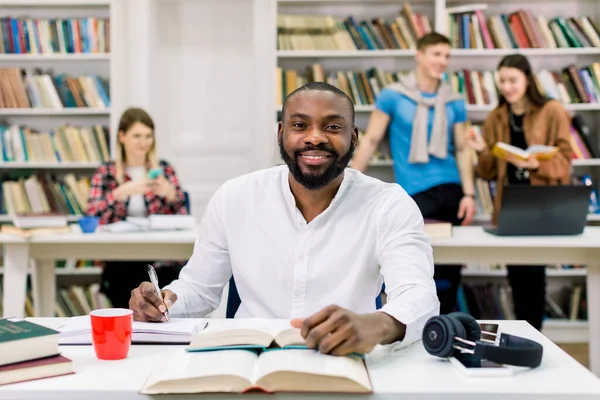 Όμορφος νεαρός χαρούμενος φιλικός Αφροαμερικάνος με γενειάδα, κοιτάζει την κάμερα και χαμογελάει, ενώ κάθεται στη βιβλιοθήκη, μελετώντας και κρατώντας σημειώσεις από το βιβλίο. Διαφορετικοί φίλοι που μιλάνε πίσω — Φωτογραφία Αρχείου