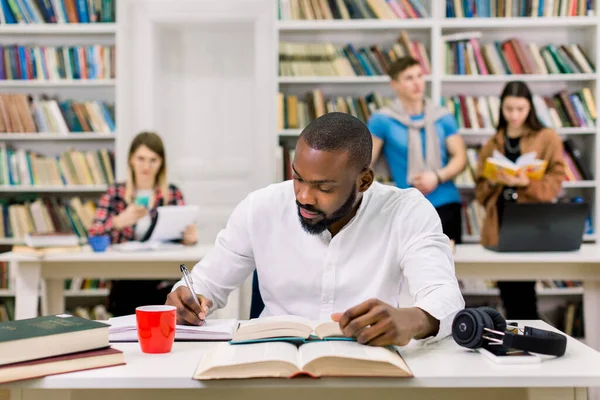 Έξυπνος ακόλουθος νεαρός Αφροαμερικάνος φοιτητής με περιστασιακή ενδυμασία που σπουδάζει στη βιβλιοθήκη του πανεπιστημίου και σημειώνει στο βιβλίο αντιγραφή, προετοιμάζεται για εξετάσεις ή εξετάσεις με τους συμπαίκτες του στο backround — Φωτογραφία Αρχείου