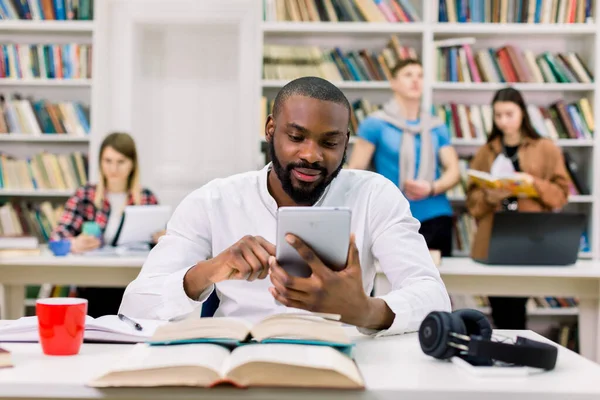 Joven hombre feliz africano guapo, estudiante, con barba y camisa blanca, sentado en la mesa con libros en la sala de lectura de la biblioteca y el uso de tableta PC, en busca de información para el estudio —  Fotos de Stock
