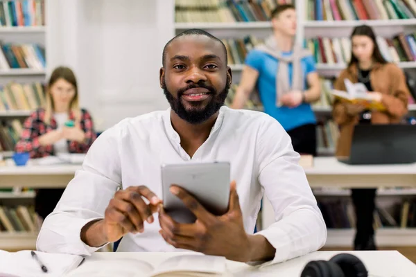 Ευτυχισμένοι νεαροί πολυφυλετικοί φοιτητές πανεπιστημίου που σπουδάζουν με βιβλία στη βιβλιοθήκη. Όμορφος αφρικανός που κοιτάζει την κάμερα ενώ κάθεται στο τραπέζι και εργάζεται σε tablet pc, i-pad — Φωτογραφία Αρχείου