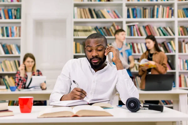 Νεαρός πολυάσχολος Αφρικανός με λευκό πουκάμισο, φοιτητής που σπουδάζει στη βιβλιοθήκη του πανεπιστημίου, μιλάει στο smartphone και κρατάει σημειώσεις στο βιβλίο του. Διαφορετικοί μαθητές που μελετούν μαζί στο παρασκήνιο — Φωτογραφία Αρχείου